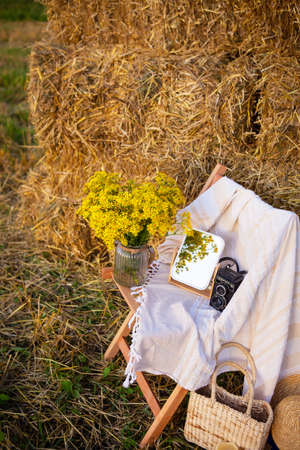 Picnic in the field near straw bales. The setting sun. Rustic style - wood chair, plaid, bouquet of flowers, mirror, hat, camera. Romantic dateの写真素材