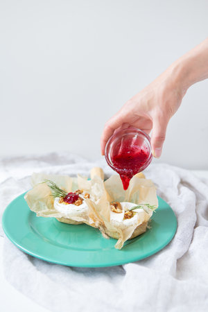 Baked camembert with walnuts, honey and rosemary wrapped in parchment lies on a green plate, a girl pours cranberry jam into the cheese, delicious and healthy foodの写真素材