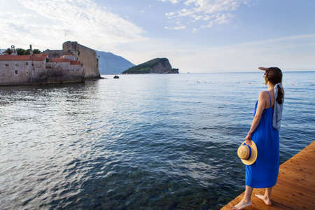 A girl in a blue dress with a straw hat stands on a wooden pier near the sea. In the background is the old medieval town in Montenegro in Budvaの写真素材
