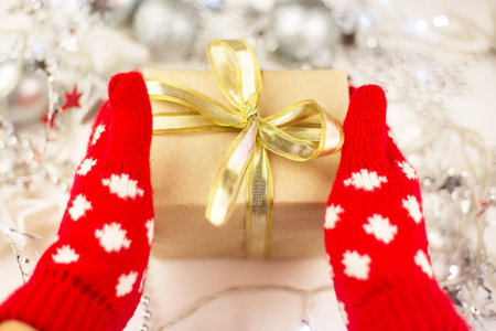Female hands hold a Christmas gift with a gold ribbon in red mittens, new year mood, preparation for the celebrationの写真素材
