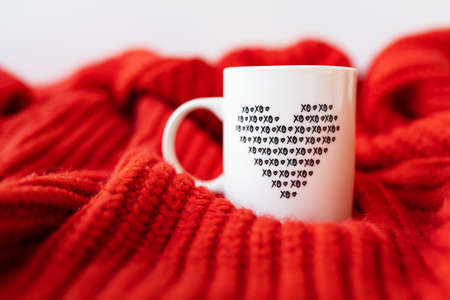 A white cup with a heart stands on a red knitted fabric. The concept of love and celebration of St. Valentine's day, February 14thの写真素材