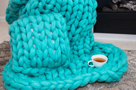 A warm plaid knitted from merino wool of a beautiful aquamarine shade lies near the fireplace along with a cup of tea. The concept of comfortの写真素材