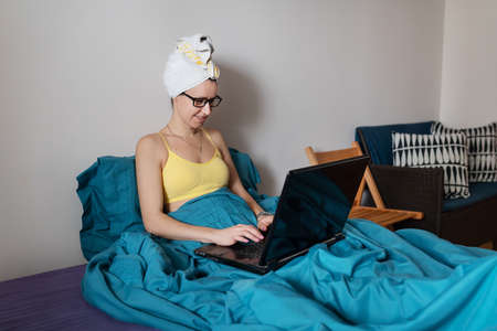 Home office, telecommuting, isolation, quarantine. A young girl working on a laptop, sitting on a bed at home in a towel after a showerの写真素材
