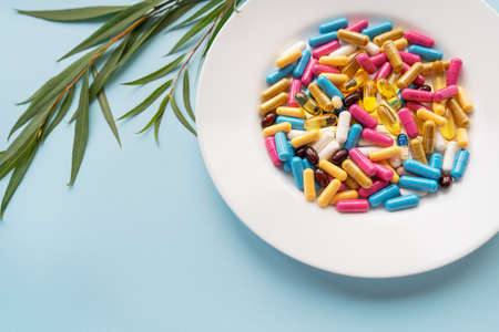 White plate with pills of nutritional supplements in different bright colors. Blue background, place for an inscription. Green branch on the backgroundの写真素材