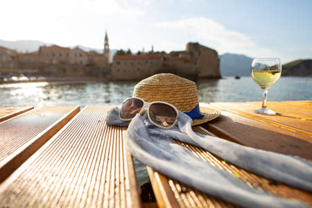 A straw hat, glasses and a glass of wine stands on a pier near the sea, close-up. Beautiful light from the sun. Place for textの写真素材