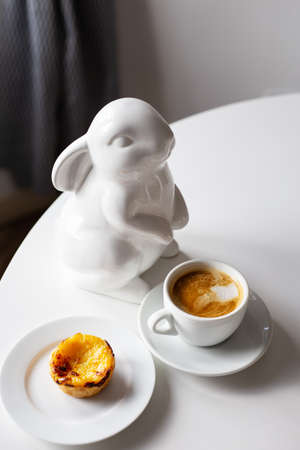 Easter Bunny stands near a cup of cappuccino and a cake on a plate, a white cup of coffee. Easter table decoration. copy spaceの写真素材