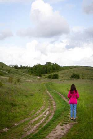 A woman in a pink raincoat looks at a mountain road. The concept of hiking in the mountains,...