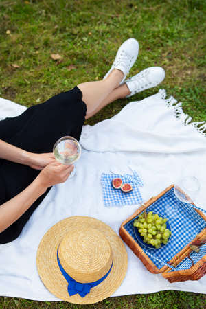 Romantic picnic in the park on the grass, delicious food: basket, wine, grapes, figs, cheese, blue checkered tablecloth, two glasses of wine. The girl is holding a glass of wine in her hands.The concept of outdoor recreationの写真素材