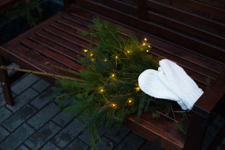 Branches of a coniferous tree decorated with a garland lie on a bench along with knitted white mittensの写真素材