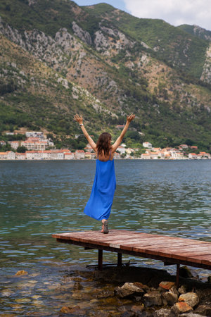 A girl in a blue dress stands with her arms raised on a pier against the background of the Bay of Kotor in Montenegro. A beautiful and cozy old townの写真素材