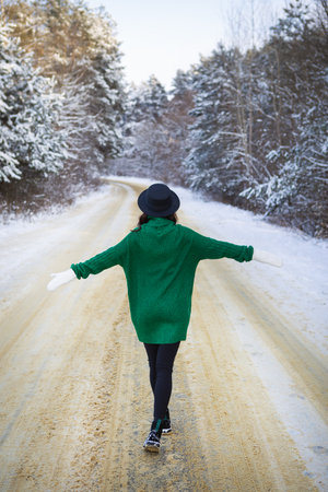 A young girl in a green sweater and hat walks in the middle of a snowy road in a thick pine forest. Freezing dayの写真素材