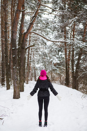 A girl in a pink scarf and hat stands with her back in the middle of a pine forest. Beautiful pine forest on a frosty dayの写真素材