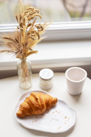 A white plate on which there is a croissant and a glass of clay with an inscription, handmade are near the window on the tableの写真素材