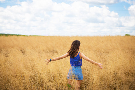 Portrait of a young beautiful brunette woman in a blue T-shirt and denim shorts stands in the middle of the field, exposing her face to the sun on the background of a summer day outdoorsの写真素材