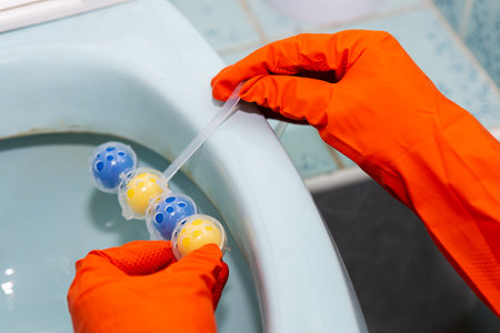 A yellow-blue plastic toilet block that cleans the walls of the toilet and refreshes the bathroom. A girl in latex red gloves hangs a toilet blockの写真素材