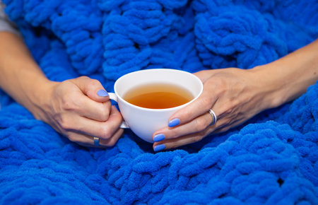 A girl wrapped in a large blue merino wool hand knitted blanket, a warm and very soft blanket, a girl holding a white cup of hot tea, place for textの写真素材