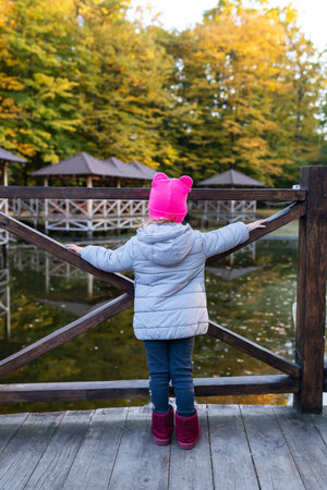 A little girl in a pink hat stands with her back near the pond and enjoys a warm autumn dayのeditorial素材