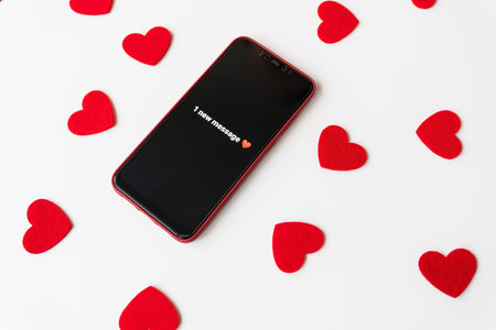 A smartphone with an activated screen with an SMS message in the form of a heart lies on a white table against a background of small red hearts. Business technology and social lifestyle concept. Valentine's Day is February 14の写真素材