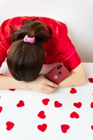 The girl is sad, puts her head in her hands, holds a smartphone in a cherry case with a heart, waiting for a text message or an invitation to a date. Valentine's day and dating conceptの写真素材