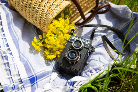 A straw picnic basket lies on a blue blanket on green grass along with a bouquet of yellow flowers. In the background is an old camera with the name Lover 166 written on itの写真素材