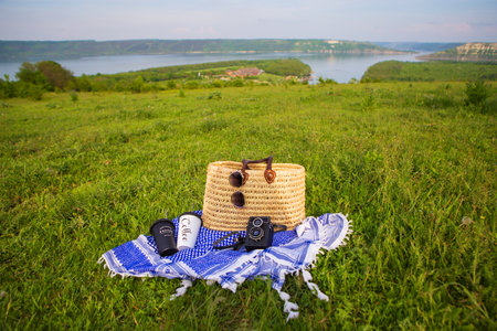 A very beautiful view of the lake. A straw picnic basket stands on a blue blanket on green grass along with glasses, a camera and coffee cups. Recreation in the open airの写真素材