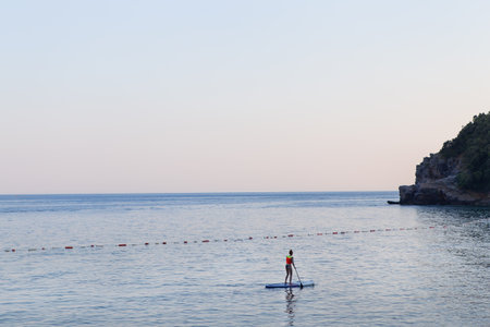 A girl with a paddle in the sea stands on a sapboard at sunrise. Active rest on the seaの写真素材