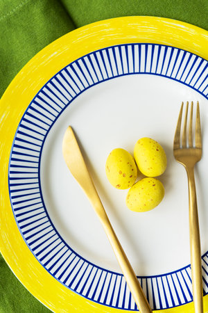 Easter serving concept. Close-up, vertical photo. Festive table setting with bright napkins and yellow-blue plates with dishes and decorated eggsの写真素材
