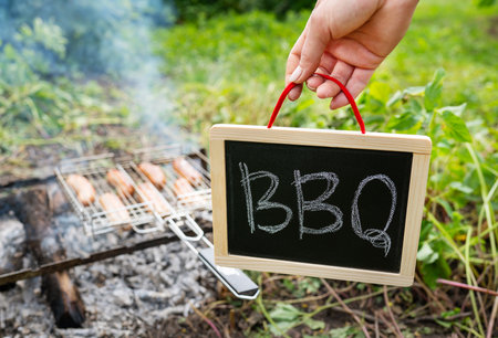 Close-up of fried sausages on the grill. A man is holding a sign with the inscription barbecue. Rest in nature, picnic, weekendの写真素材