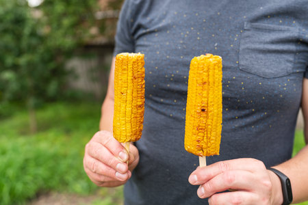 In his hands, a man holds roasted sweet corn with salt and butter on a stick on a grill on the background of a recreation park. Summer vegan dinner or appetizer, grillingの写真素材