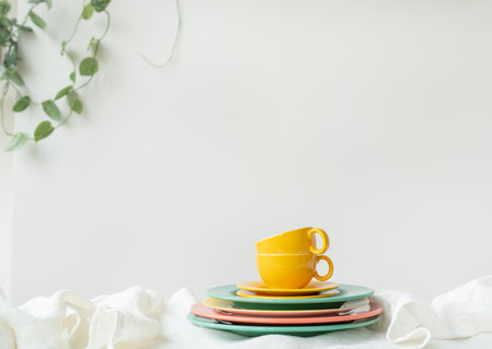 Empty yellow cups and brightly colored plates on a white table covered with a linen tablecloth, a flower pot on the wall in the background. preparation for servingの写真素材