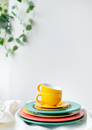 Empty yellow cups and brightly colored plates on a white table covered with a linen tablecloth, a flower pot on the wall in the background. vertical photoの写真素材