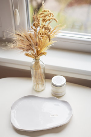 A white plate made of handmade clay, a candle, a vase stands near the windowの写真素材