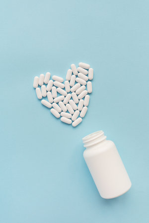 White pills in the shape of a heart came out of a jar on a blue background, health and heart problems together with a plastic jar. The concept of medicine and health care. Place for writingの写真素材