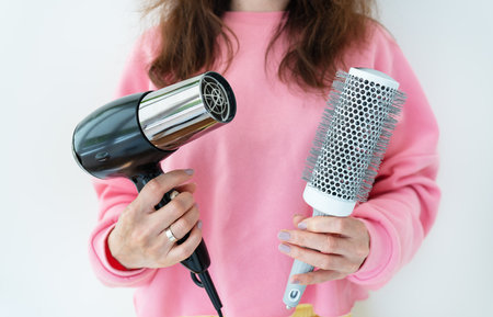 The girl holds a hair dryer and a brush in her hands. Drying long brown hair with a round brush for styling unruly hair. beauty saloonの写真素材