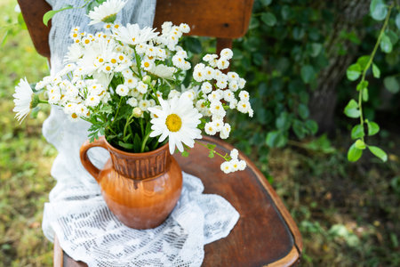 Summer atmosphere, simple home decor in the countryside. Beautiful white daisies on a wooden old chair along with a lace napkin against the backdrop of a green gardenの写真素材