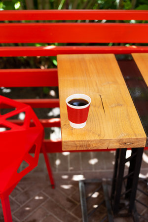 A wooden table on which there is a red plastic cup with coffee. Summer beautiful terrace in greeneryの写真素材
