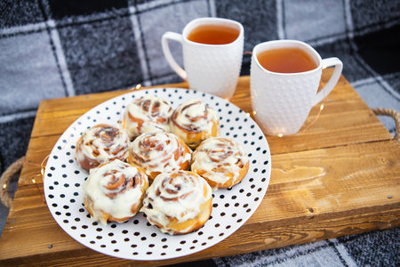 Two cups of black tea stand on a wooden tray on the sofa with a black and white checkered plaid. Fresh and fragrant cinnamon rolls close up lie on a plate with polka dots, beautiful morning.の写真素材
