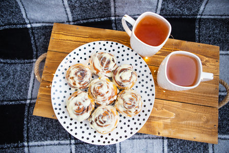 Two cups of black tea stand on a wooden tray on the sofa with a black and white checkered plaid. Fresh and fragrant cinnamon rolls close-up lie on a plate with polka dots. The garland is blinking.の写真素材