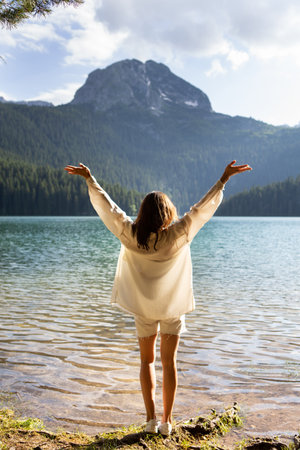 The girl stands with her back near a mountain lake in the background. Positive young woman traveling on a blue lake outdoors, adventure travelの写真素材