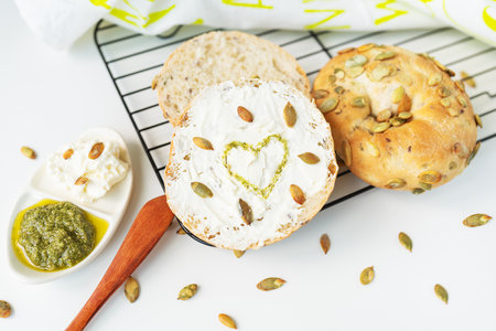 A freshly baked bagel with Philadelphia cheese, sunflower seeds scattered on the table and a heart painted with pesto sauce. Delicious and healthy breakfastの写真素材