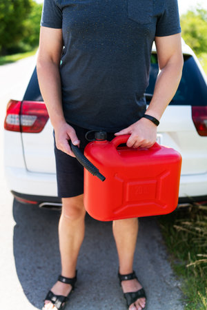 A man is holding a red can next to a white car. Stop to refuel, problems on the road with refueling, self-refueling. Vertical photoの写真素材