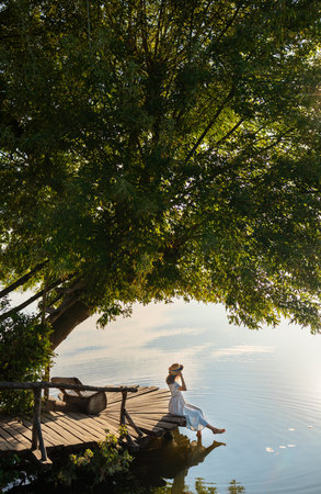 A young beautiful girl in a hat and a long dress sits on a pier by the lake and looks at the rising sun. The concept of travel and outdoor recreationの写真素材