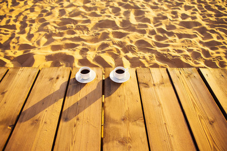 two cups of coffee stand on a pier near a sandy beach.の写真素材