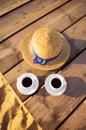 A hat and two cups of coffee stand on a pier near a sandy beach.の写真素材