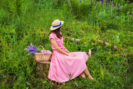 beautiful girl in a beautiful dress sits on a stump in the forest, a bouquet of lupines in a basket.の写真素材