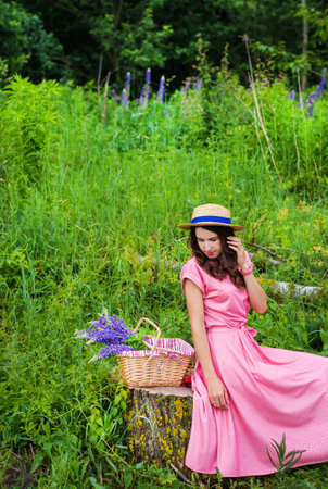 beautiful girl in a beautiful dress sits on a stump in the forest, a bouquet of lupines in a basket.の写真素材