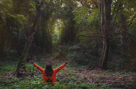 A girl in an orange sweater with a scary face from the Halloween holiday sits in a dense dark autumn forest. Halloween holiday October 31stの写真素材