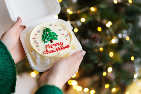 A girl holds in her hands a homemade Christmas cake with the inscription Merry Christmas, against the background of a decorated Christmas treeの写真素材