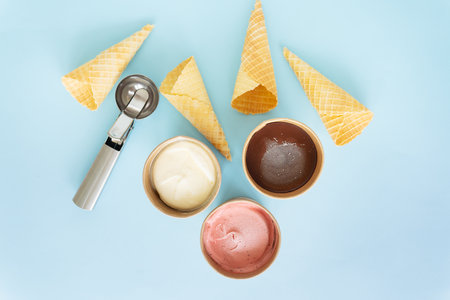 Ice cream in a box with different flavors stands on a blue background along with a metal ice cream scoop and a waffle cone. Selective focus, noiseの写真素材
