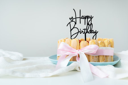 Delicious and bright strawberry tiramisu cake with cheese cream, decorated with a bow, stands on the table, the inscription Happy Birthday is on top, a place for an inscriptionの写真素材